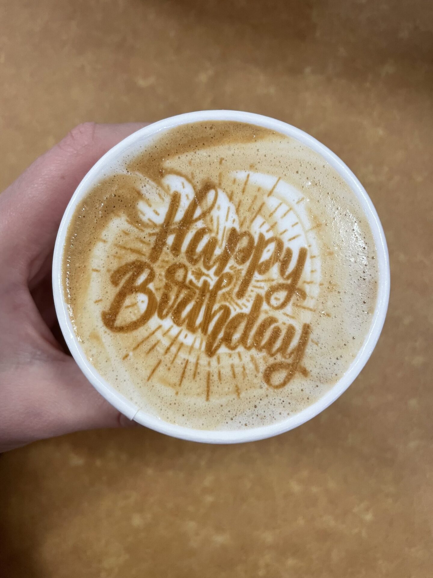 A hand holding a coffee cup with the words "happy birthday" on the foam