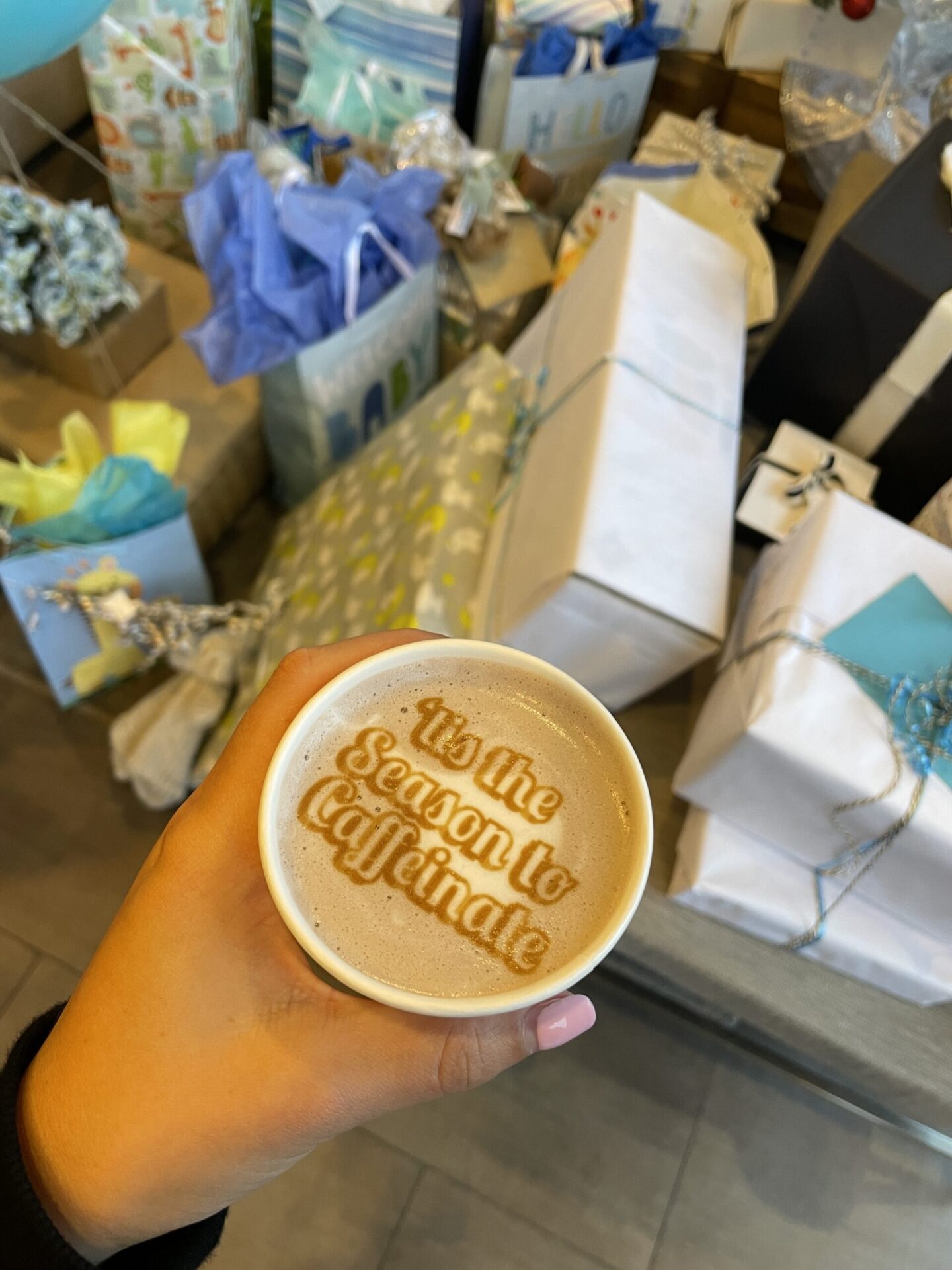 A hand folding a coffee cup with the words "'tis the Season to Caffeinate" on the foam in front of christmas presents